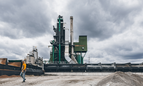 Asfaltcentrale met groene en grijze installaties onder bewolkte hemel. Op de voorgrond loopt een arbeider in oranje veiligheidskleding langs zwarte containers.