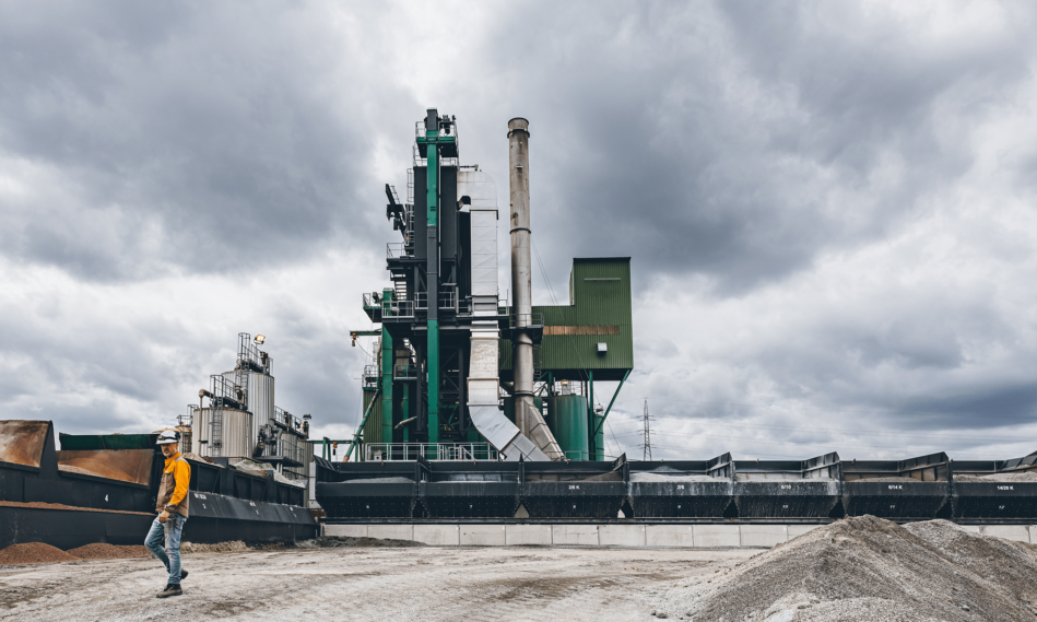 Asfaltcentrale met groene en grijze installaties onder bewolkte hemel. Op de voorgrond loopt een arbeider in oranje veiligheidskleding langs zwarte containers.